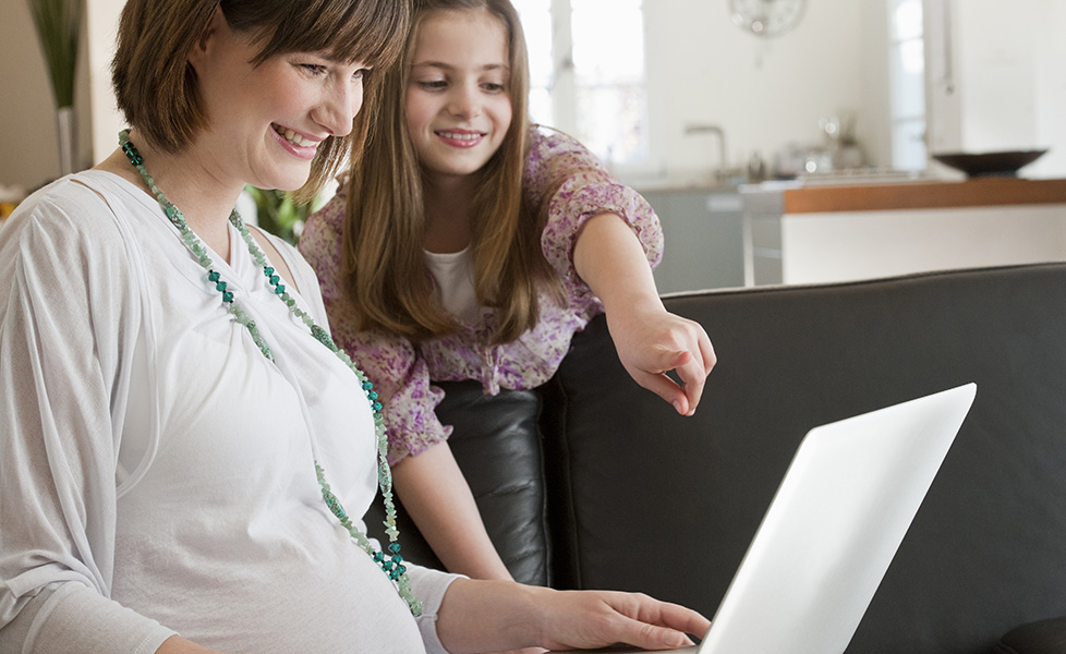 Pregnant mid adult mother and daughter using laptop on sofa